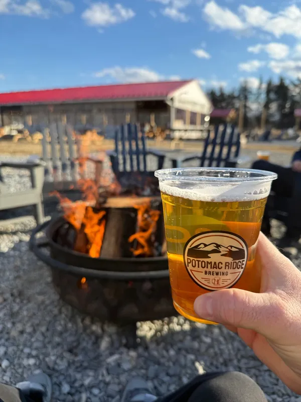 A cup of golden beer with a Potomac Ridge Brewing logo held up in front of an outdoor fire pit, Adirondack chairs and a red-roofed building
